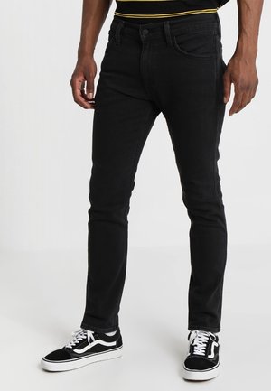 Jeans neri slim fit indossati con sneakers nere e bianche e una maglietta nera con strisce gialle in vita.