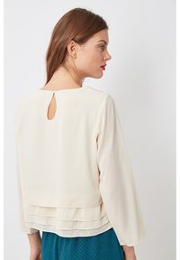 Blusa blanca de manga larga, con un detalle de abertura en la espalda y volantes en el dobladillo, confeccionada en una tela suave.