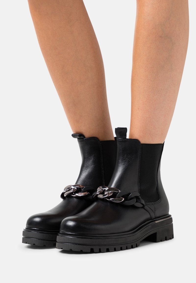 Bottines noires en cuir et élastique avec une semelle épaisse. Présentent une chaîne métallique décorative à l'avant et une languette de traction.