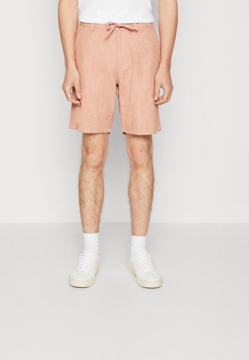 Homme portant un short rose clair à cordon, des chaussettes blanches et des baskets blanches, debout devant un fond clair uni.