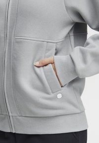 Light gray zip-up hoodie made of soft fabric, featuring front pockets and ribbed cuffs. Includes a small metal accent detail.