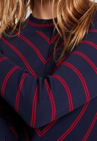 Maglione blu navy con strisce orizzontali rosse, texture a maglia e colletto a coste. Dettaglio in primo piano della manica e del braccio incrociato.