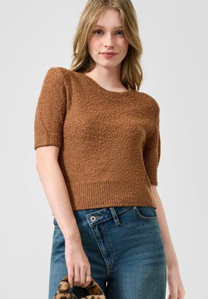 Femme portant un pull marron texturé à manches courtes et un jean bleu, tenant un petit sac avec une poignée imprimée léopard.