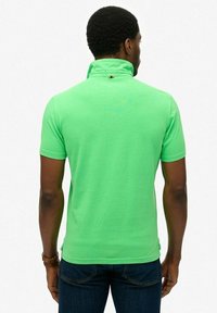 Zielona koszulka polo z podniesionym kołnierzykiem i krótkimi rękawami, wykonana z gładkiego materiału. Widoczny detal z logo z tyłu, blisko kołnierzyka.