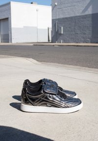 Par de zapatillas negras brillantes con suelas blancas y cordones negros colocadas sobre una acera de hormigón en una zona urbana.