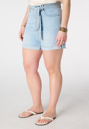 Femme portant un short en denim bleu clair avec une ceinture nouée, un haut blanc, plusieurs bracelets, des bagues, et des tongs blanches à perles, debout sur un fond uni.