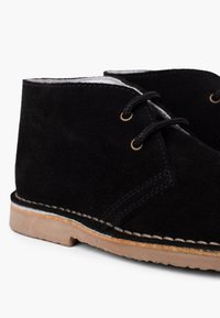 Botas de tobillo de ante negro con punta redonda, que presentan cordones negros y ojales dorados, acentuadas con costuras beige y una suela en tono marrón claro.