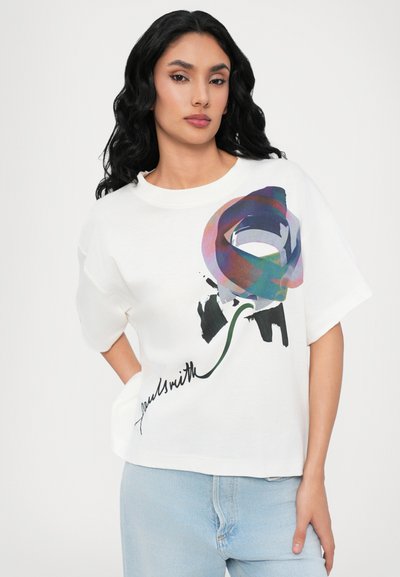 Hvit overdimensjonert t-skjorte med et flerfarget abstrakt mønster og svarte detaljer, kombinert med lyseblå jeans. Korte ermer og rund hals.