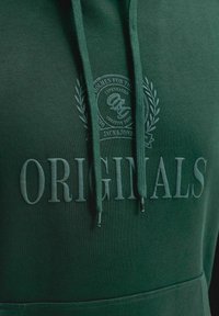 Σκούφος πράσινος με ανάγλυφο κείμενο "ORIGINALS" και κυκλικό έμβλημα που φιλοξενεί "Jack & Jones" και φύλλα δάφνης στο στήθος.