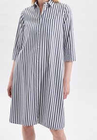 Striped button-up shirt dress in white and navy, featuring a collar, three-quarter sleeves, and a chest pocket. Lightweight fabric, relaxed fit.