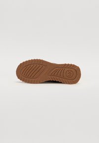 Semelle en caoutchouc marron avec un motif de grip texturé et un détail de logo circulaire, présentant des rainures et des stries pour une traction et un support.