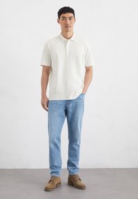 Herren Poloshirt in Creme mit kurzen Ärmeln, versehen mit einem kleinen bestickten Logo, kombiniert mit hellblauen Jeans und braunen Wildlederschuhen.