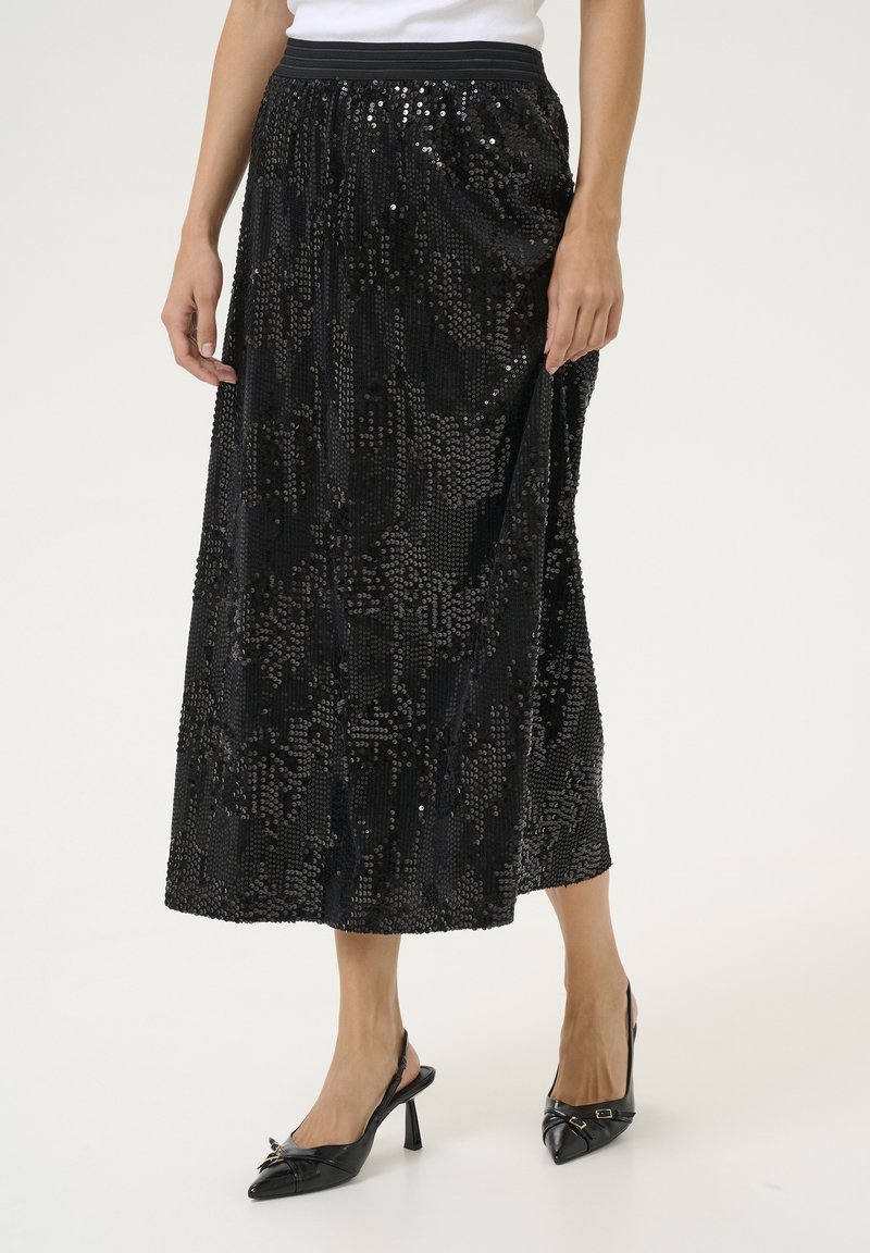 Femme portant une jupe midi noire à sequins avec une ceinture élastique noire et des talons slingback noirs à bouts pointus avec de petits nœuds.