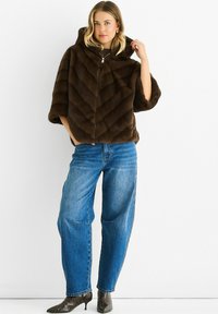 Woman wearing a brown fur zip-up hooded jacket, blue high-waisted jeans, and snakeskin-patterned pointed boots, standing against white wall.