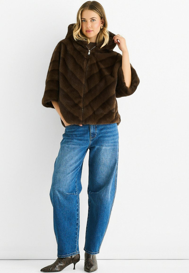 Woman wearing a brown fur zip-up hooded jacket, blue high-waisted jeans, and snakeskin-patterned pointed boots, standing against white wall.