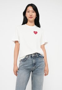 Bela bombažna majica s rdečim srčnim grafikom, na kateri je napisano "Upanje" v kurzivi. V kombinaciji s pobledelimi modrimi jeans hlačami.