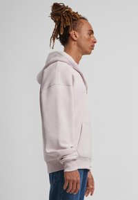 Felpa rosa chiaro con tasca frontale, cappuccio con coulisse e polsini a costine. Presenta una consistenza liscia e una vestibilità comoda.