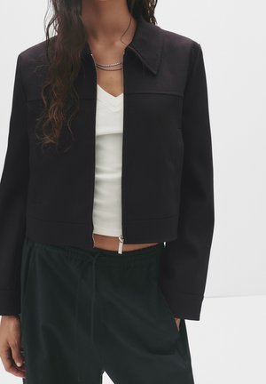 Femme portant une veste noire courte zippée par-dessus un haut blanc à col en V et un pantalon sombre à cordon de serrage, debout devant un fond uni.