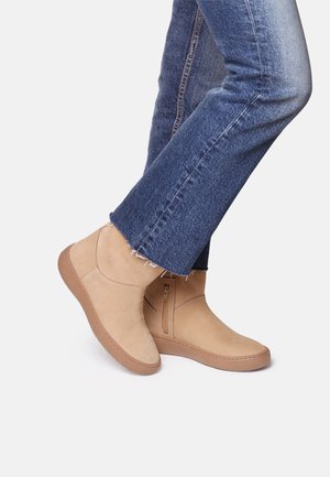 Bottines en daim beige à fermeture éclair latérale, associées à un jean en denim bleu légèrement effiloché et évasé, présentées dans une position de marche.