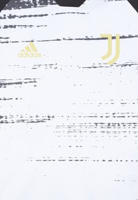 Weißes Kurzarmhemd mit einem strukturierten schwarzen Streifenmuster. Enthält das Adidas-Logo in Gold und das Juventus-Logo auf der rechten Seite.