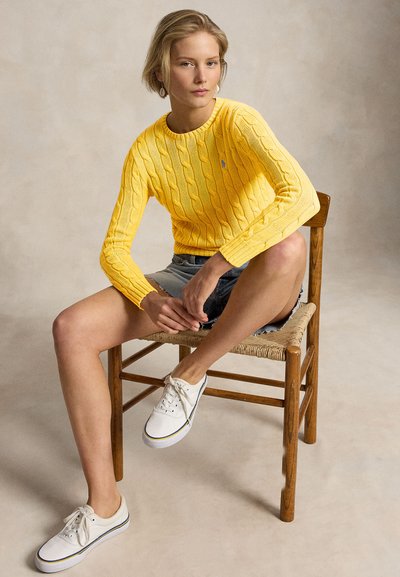 Νεαρή γυναίκα κάθεται σε ξύλινη καρέκλα φορώντας κίτρινο πουλόβερ με κόμπους, τζιν σορτς και λευκά αθλητικά παπούτσια μπροστά σε ουδέτερο φόντο.