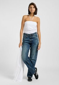 Weißes, trägerloses Oberteil mit Lochmuster, kombiniert mit lässigen dunkelblauen Jeans. Schwarze Sneakers mit weißen Streifen und ein weißes Hemd in der Hand.