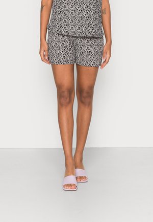 Personne portant un short à fleurs noir et blanc, un haut assorti, et des sandales slide ouvertes à l'avant de couleur rose pâle, debout devant un fond uni.