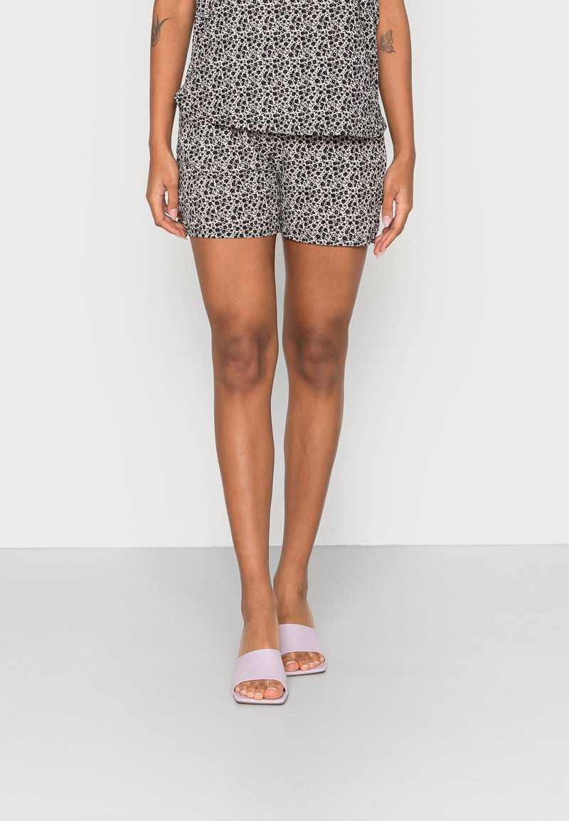 Personne portant un short à fleurs noir et blanc, un haut assorti, et des sandales slide ouvertes à l'avant de couleur rose pâle, debout devant un fond uni.