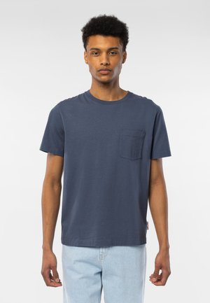 Ung mann med kort krøllete hår som har på seg en ensfarget marineblå t-skjorte med korte ermer og brystlomme og lyseblå jeans, stående mot en hvit bakgrunn.
