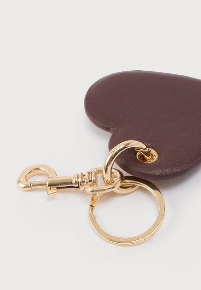 Καφετί μπρελόκ σε σχήμα καρδιάς από λείο υλικό, με χρυσό μεταλλικό κούμπωμα και δαχτυλίδι για να το κρεμάτε σε κλειδιά ή τσάντες.