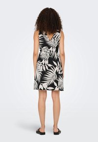 Femme aux cheveux bouclés portant une robe sans manches noir et blanc à motif de feuilles, debout pieds nus sur un fond uni, de dos.