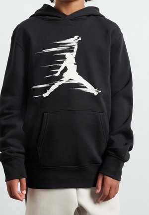 Enfant portant un sweat à capuche noir avec une poche avant et un graphisme blanc stylisé d'une silhouette en course sur la poitrine, associé à un pantalon clair.