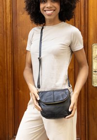 Borsa a tracolla in pelle nera con pattina curvata e chiusura a zip, dotata di una tracolla sottile e accessori in metallo dorato. Texture morbida e dimensioni compatte.