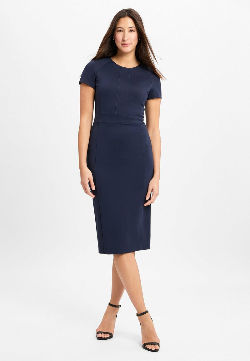 Navyblaues, tailliertes Kleid mit kurzen Ärmeln, rundem Ausschnitt und Nahtdetails. Getragen mit schwarzen Ankle-Strap-High Heels. Glattes, elastisches Material.