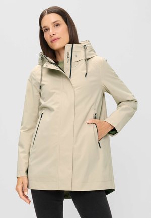 Frau trägt beige Kapuzen-Regenmantel mit Reißverschlusstaschen, steht mit einer Hand in der Tasche vor hellgrauem Hintergrund.