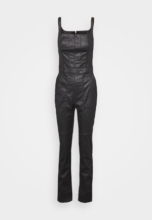 Sort ærmeløs jumpsuit i kunstlæder med firkantet halsudskæring, tætsiddende talje og bukser med lige ben på ensfarvet baggrund.