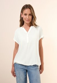 Blusa blanca ligera con escote en V, mangas cortas enrolladas y botón en la parte delantera. Textura suave con un corte holgado y dobladillo fruncido.