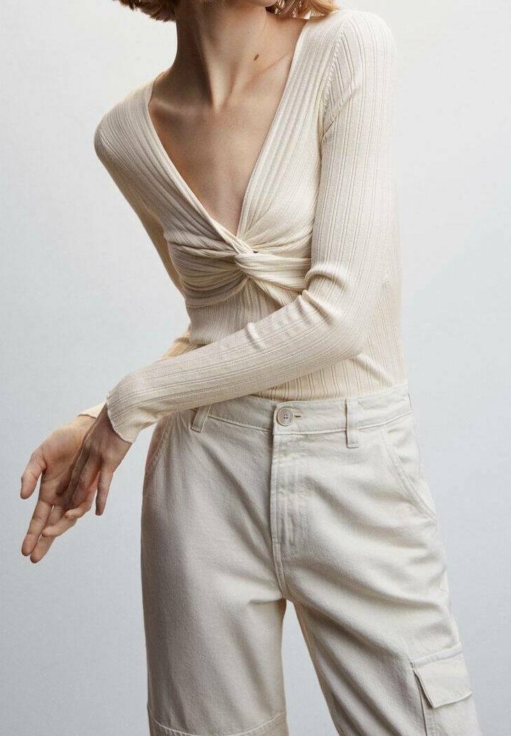 Femme portant un top à manches longues côtelé couleur crème avec un nœud torsadé au niveau du col en V, associé à un pantalon cargo taille haute beige clair.