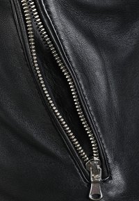 Närbild av svart läder med en metallisk dragkedja med synliga tänder och en silverfärgad dragflik, mot en bakgrund av texturerat tyg.