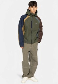 Meerlagig lichtgewicht jack met capuchon in meerdere kleuren; met groene, blauwe, bruine en beige delen; ritssluiting; gladde textuur; gelaagd ontwerp.