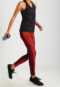 Czarny sportowy top na ramiączkach w połączeniu z bordowymi legginsami z czerwonym zygzakowatym wzorem; model trzyma butelkę wody, nosząc czarne sneakersy.