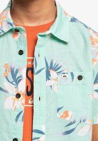 Kurzärmliges Button-Down-Hemd in Minzgrün mit floralen Muster in Orange, Pink und Blau; hat braune Knöpfe und eine Brusttasche.