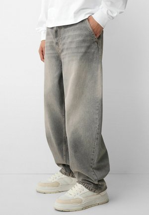 Personne portant un jean gris délavé à coupe ample avec des poignets élastiques et des baskets blanches, debout avec une main dans la poche.