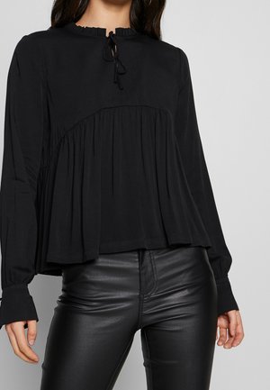 Kobieta nosząca czarną bluzkę z długim rękawem, marszczonym dekoltem i luźnym krojem, w połączeniu z czarnymi skórzanymi spodniami, na jednolitym tle.