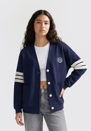 Kvinde med langt mørkt hår iført navy cardigan med hvide striber på ærmerne og emblem, hvid crop top og grå jeans, stående mod hvid baggrund.
