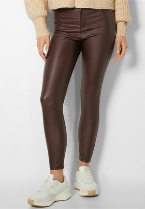 Person iført tætsiddende mørkebrune leggings med læderlook, beige strikket sweater og hvide chunky sneakers, stående mod en ensfarvet baggrund.