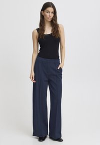 Femeie stând cu o mână în buzunar, purtând un top negru fără mâneci și pantaloni largi bleumarin, pe un fundal simplu, deschis.