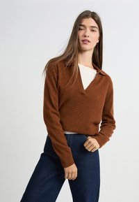 Sweter w kolorze rdzy z dekoltem w kształcie litery V i kołnierzykiem, z długimi rękawami i luźnym krojem. Noszony na jasnej koszuli, w zestawieniu z ciemnymi dżinsami.