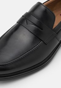 Mocassin noir en cuir sans lacets avec détail de lanière penny et accents cousus, doté d'un bout carré arrondi et d'une finition lisse.