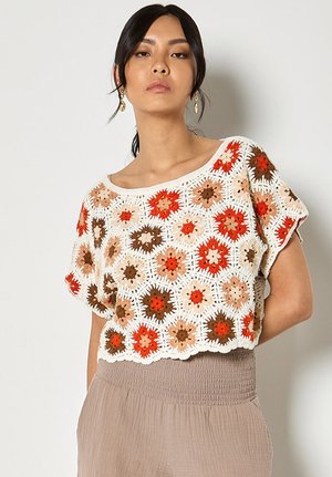 Frau trägt ein kurz geschnittenes Häkeloberteil mit mehrfarbigen sechseckigen Blumenmustern in Orange, Braun und Beige, kombiniert mit beigen, hoch taillierten Hosen.